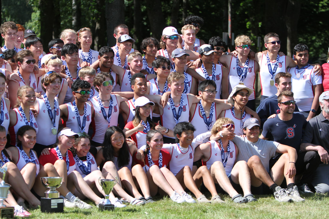 The Sammamish Rowing Association junior team all stands together for a team photo including coaches. The sky is blue and grass is green. 